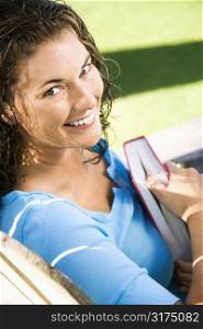 Portrait of smiling pretty young adult Caucasian brunette female sitting in chair holding book and looking at viewer.