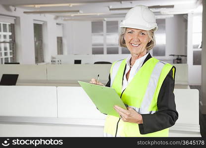 Portrait of smiling senior woman in reflector vest and hard hat holding clipboard at office