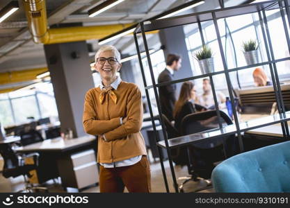 Portrait of smiling senior woman standing outside meeting room with team discussing work in background