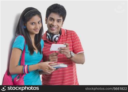 Portrait of smiling university friends with mobile phone and books against wall