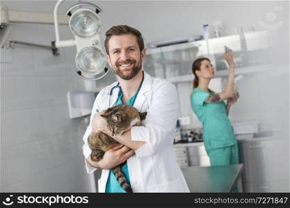 Portrait of smiling veterinary doctor carrying cat while standing against coworker at clinic