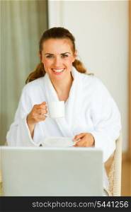Portrait of smiling woman in bathrobe with cup of coffee and laptop on table