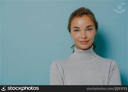 Portrait of smiling woman with makeup and healthy glowing skin dressed in grey poloneck looks directly at camera against blue background copy space for your advertisement. Human face expressions. Portrait of smiling woman with makeup and healthy glowing skin dressed in grey poloneck