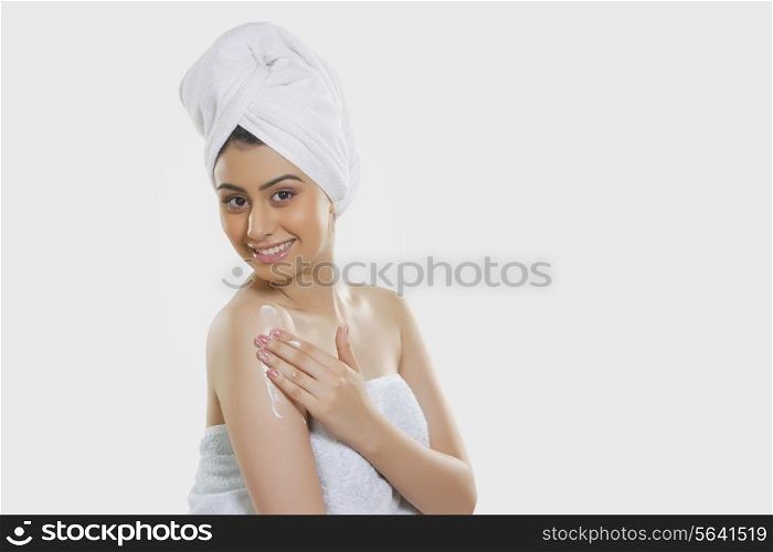 Portrait of smiling woman wrapped in towel applying moisturizer on arm over white background