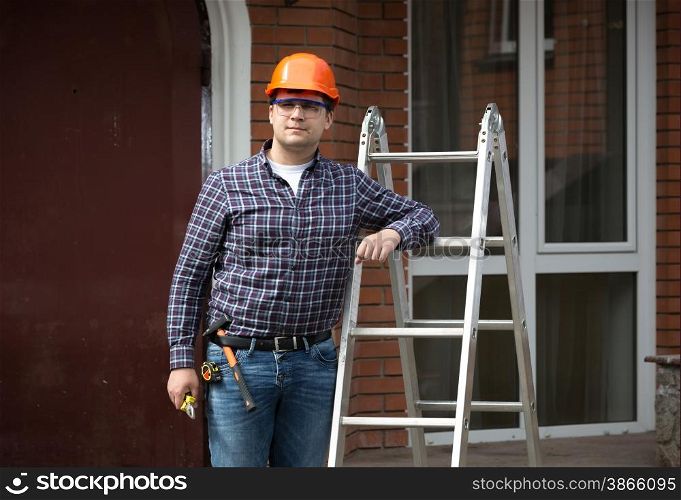 Portrait of smiling worker in hard hat leaning against metal ladder