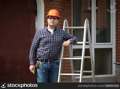 Portrait of smiling worker in hard hat leaning against metal ladder