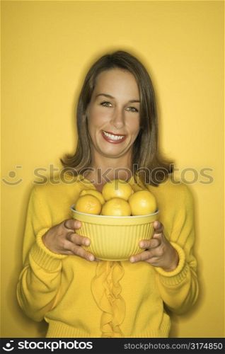Portrait of smiling young adult Caucasian woman on yellow background holding bowl of lemons.