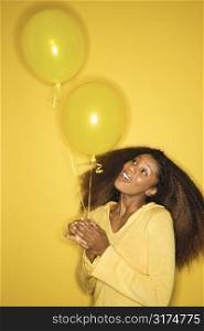 Portrait of smiling young African-American adult woman on yellow background holding balloons.