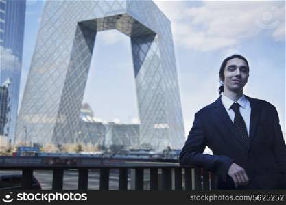 Portrait of smiling young businessman leaning on the railing with the CCTV building in background, Beijing