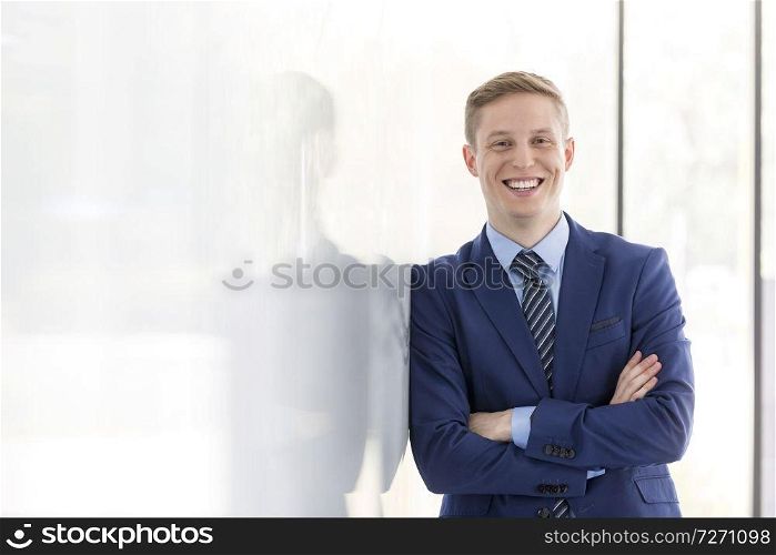 Portrait of smiling young businessman with arms crossed leaning on wall at office