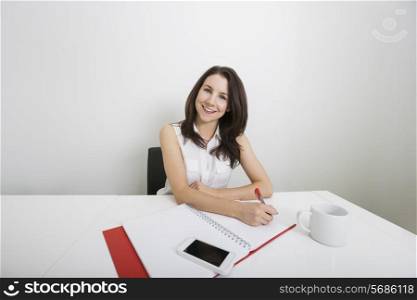 Portrait of smiling young businesswoman writing on binder at desk in office