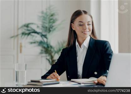 Portrait of smiling young caucasian bussiness woman in black formal suit sitting by big white desk working with laptop and writing things down with blurred light background. People at work concept. Happy bussiness woman working on laptop at office