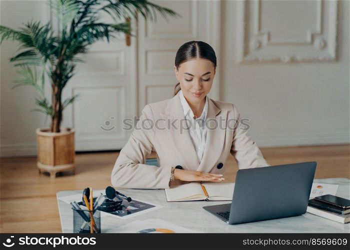 Portrait of smiling young caucasian bussiness woman in light beige formal suit with hair tied in ponytail, sitting by big white desk working with laptop and writing things down with blurred background. Concentrated bussiness woman sitting by desk in front of laptop
