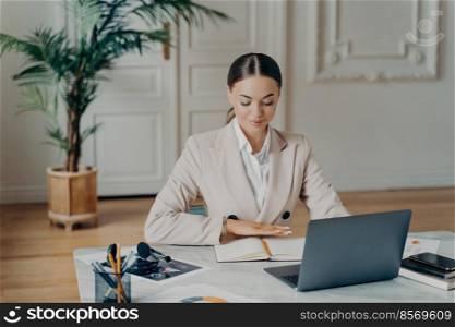 Portrait of smiling young caucasian bussiness woman in light beige formal suit with hair tied in ponytail, sitting by big white desk working with laptop and writing things down with blurred background. Concentrated bussiness woman sitting by desk in front of laptop