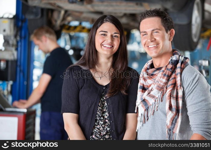 Portrait of smiling young couple in auto repair shop with mechanic in the background