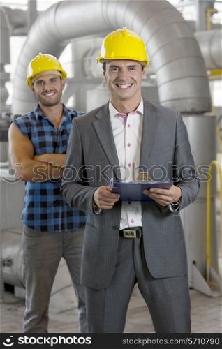 Portrait of smiling young male manager holding clipboard with worker in background at industry