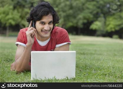 Portrait of smiling young man lying on grass while using cell phone and laptop