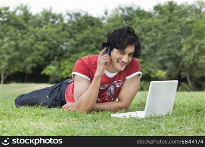 Portrait of smiling young man using cell phone and laptop while lying on grass