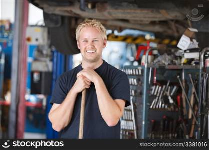 Portrait of smiling young mechanic leaning on a broom in a garage