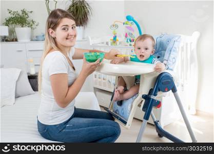 Portrait of smiling young mother feeding her toddler sin in highchair and looking in camera. Smiling young mother feeding her toddler sin in highchair and looking in camera