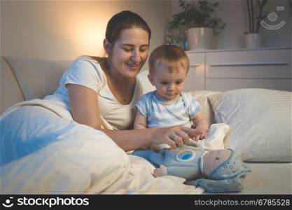 Portrait of smiling young mother playing with her baby on bed at night
