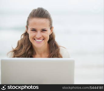 Portrait of smiling young woman in sweater sitting on beach with laptop