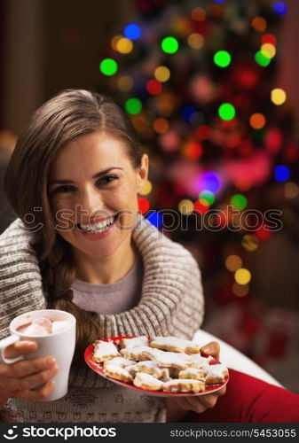 Portrait of smiling young woman with cup of hot beverage and christmas cookies near christmas tree