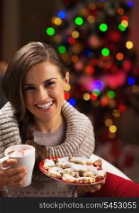 Portrait of smiling young woman with cup of hot beverage and christmas cookies near christmas tree