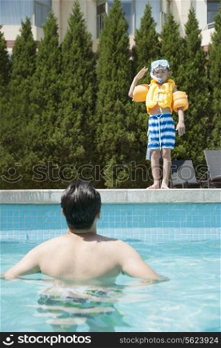 Portrait of son with snorkeling equipment standing by the pool as father waits in the water