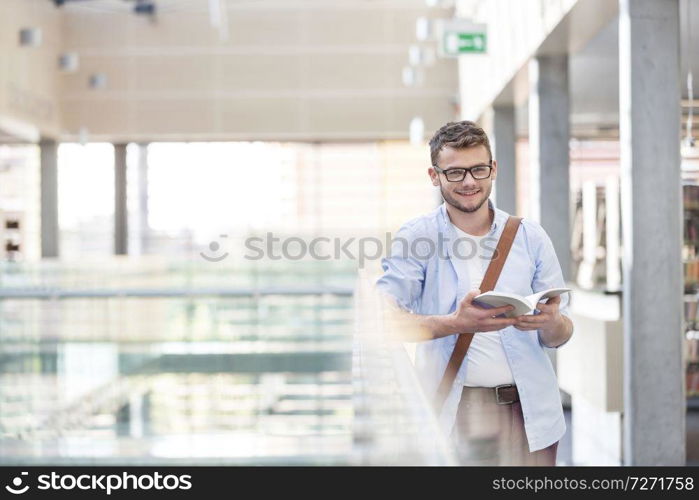 Portrait of student reading book while standing by railing at university