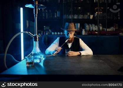 Portrait of stylish bearded man with hat smoking hookah. Fashion guy sitting at table and blowing steam vortexes. Portrait of stylish bearded man with hat smoking hookah