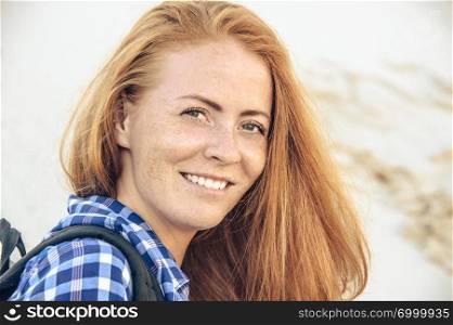 Portrait of stylish hipster young girl with red hair and freckles in a checkered shirt on slope dune. With backpack . Travel, hiking and active lifestyle concept.. Hipster young girl enjoying sun on slope dune.