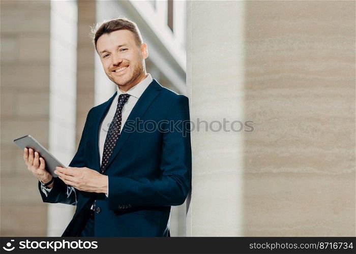 Portrait of stylish male entrepreneur has cheerful expression, holds digital tablet, reads financial news on web page, uses free internet connection, wears formal suit and tie. Career concept
