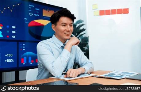 Portrait of successful confident businessman, manager or executive in business wear analyzing financial report with dashboard data on TV screen in the background at harmony office meeting room.. Portrait of male manager in harmony office with businesspeople in background.