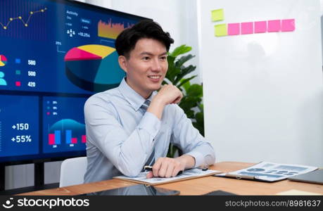 Portrait of successful confident businessman, manager or executive in business wear analyzing financial report with dashboard data on TV screen in the background at harmony office meeting room.. Portrait of male manager in harmony office with businesspeople in background.