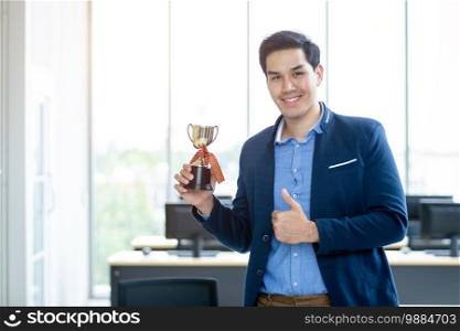 Portrait of successful feeling winner screaming handsome young asian businessman holding a ch&ion cup and showing thumbs up at In the office room background.