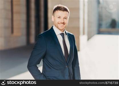 Portrait of successful intelligent businessman wears formal black suit, has positive expression, poses outdoor, rejoices successfully signed contract with business partners. Cheerful entrepreneur