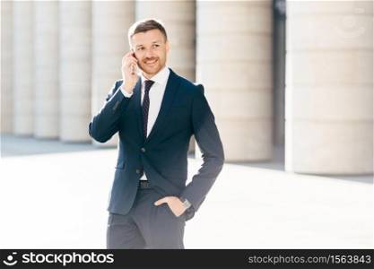 Portrait of successful male manager satisfied with mobile tariffs, makes phone call, uses banking service operator, wears formal suit, stands in urban setting outsie. Professional man worker