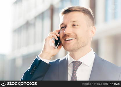 Portrait of successful positive office employee has phone conversation, looks happily into distance, discusses money payment. Male executive manager in formal suit speaks with client via cellular