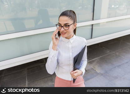 Portrait of successful young businesswoman using smartphone and holding documents outdoors.