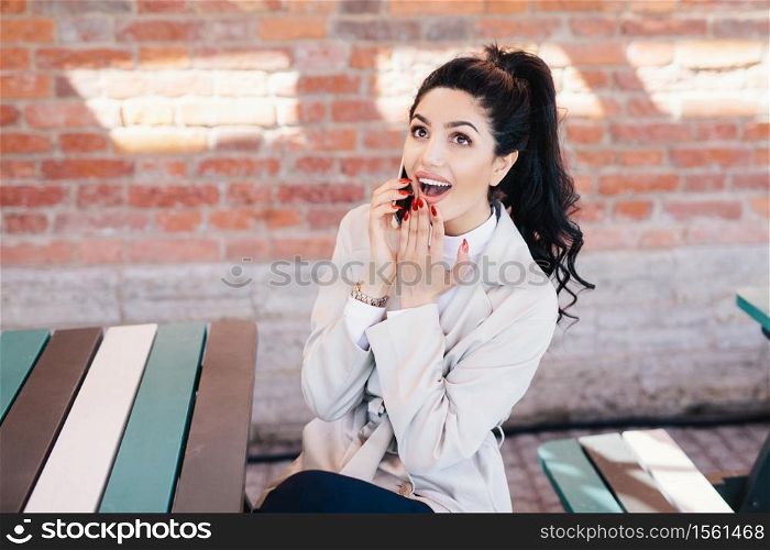 Portrait of surprised beautiful woman with dark pony tail sitting at cafe chatting with her best friend over smartphone gossiping having shocked look after hearing interesting and intriguing news