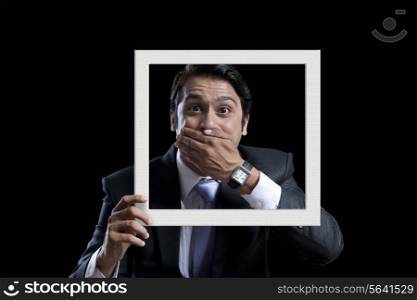 Portrait of surprised businessman covering mouth while holding frame against black background