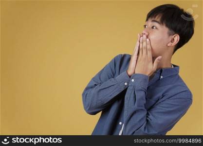 portrait of surprised excited young asian man dressed casually with hands covering mouth isolated on yellow studio background