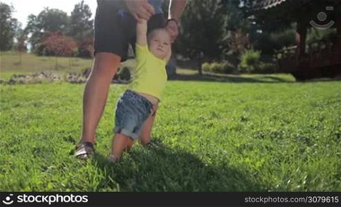 Portrait of sweet blue eyed toddler boy taking first steps with father&acute;s help on green grassy lawn in summer park. Happy little baby learning to walk with the help of his dad outdoors as they enjoy leisure together. Slow motion. Stabilized shot.