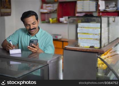 Portrait of Sweet shop owner maintaining accounts by using Smartphone at counter