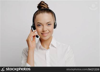 Portrait of tech support worker in classic outfit, black headset, explains problem cause, offers solution to caller. Stands isolated by light wall, looks at camera.