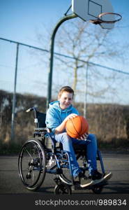 Portrait Of Teenage Boy In Wheelchair Playing Basketball On Outdoor Court