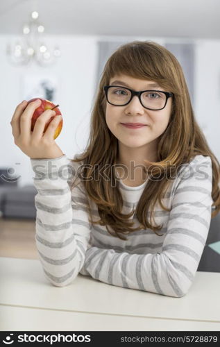 Portrait of teenage girl holding apple while sitting at table in house