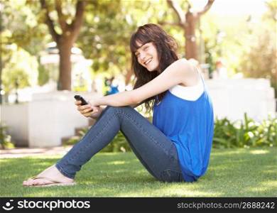 Portrait Of Teenage Girl Sitting In Park Using Mobile Phone
