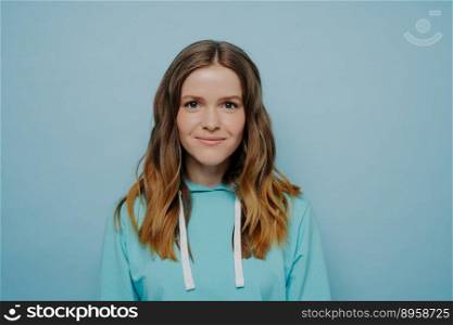 Portrait of teenager girl with wavy ombre hairstyle smiling while looking at camera in comfortable casual blue sweatshirt standing against studio background. Smiling teenage girl looking at camera posing isolated against blue studio wall